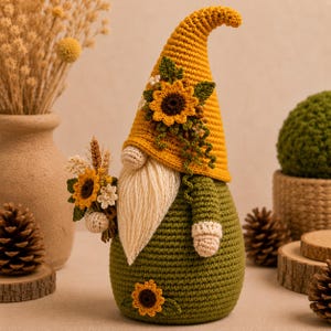 May include: A crochet gnome figurine featuring a tall yellow hat adorned with a sunflower and green leaves. The gnome has a cream-colored yarn beard, a green body with a sunflower accent, and holds a small bouquet of sunflowers and wheat.