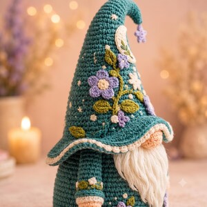 May include: A teal crochet gnome featuring a tall pointed hat decorated with embroidered lavender flowers, green leaves, a white crescent moon, and a small star at the tip. The gnome has a textured body, a cream-colored yarn beard, and light skin tone.