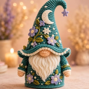 Op de afbeelding: Een gehaakte kabouter in blauwgroen met een hoge, puntige muts versierd met een crèmekleurige halve maan, een kleine paarse ster en diverse paarse en witte bloemen met groene bladeren. De kabouter heeft een lange, crèmekleurige baard van garen.