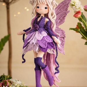 Op de afbeelding: Een handgemaakte gehaakte feeënpop met een grillig ontwerp. De pop heeft een lichte huidskleur, blond haar en een paarse en lavendelkleurige jurk met bijpassende vleugels. De pop staat op een houten voet, wat de ingewikkelde details van het haakwerk laat zien.