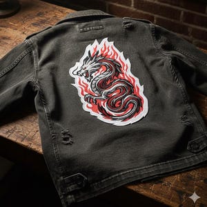 May include: Dark gray denim jacket featuring a large embroidered dragon design on the back. The dragon is red, white, and black, set against a backdrop of flames. The jacket has a distressed look with ripped details.