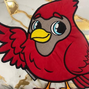 Op de afbeelding: Een rood geborduurd kardinaal-patch met een cartoonachtig ontwerp. De vogel heeft een grijs gezicht, grote blauwe ogen en een gele snavel. De patch is zwart omlijnd en voorzien van de tekst "MEHAR EMBROIDERY".