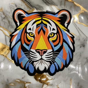 Puede incluir: Parche bordado de un tigre con un diseño colorido. La cara del tigre presenta detalles en naranja, amarillo, azul y blanco, con un contorno negro. El parche tiene el texto "HENNA EMBROIDERY". El fondo es un patrón de mármol.