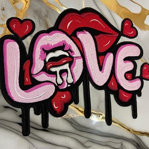 May include: Embroidered patch featuring the word "LOVE" in a dripping font, with red and pink lips and heart accents. The patch has a black border and is set against a marble background.