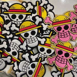 May include: Embroidered patches featuring a skull and crossbones design with a straw hat. The patches are in white, yellow, red, and black. Some patches are pink and yellow. The patches are likely for clothing or accessories.