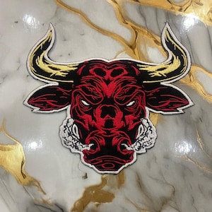 May include: Embroidered patch featuring a red bull's head with black and white accents. The bull has yellow horns and an angry expression. The patch is likely for clothing or accessories.