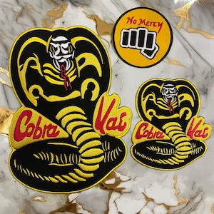 以下が含まれることがあります： Cobra Kaiのロゴと赤い文字で「Cobra Kai」と書かれた刺繍パッチ。パッチには、黒と黄色の大きなコブラの頭のデザインと、「No Mercy」と拳が描かれた円形のパッチが含まれています。