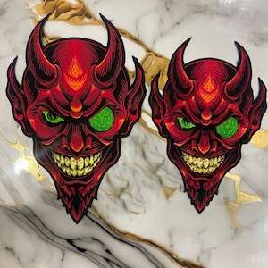 May include: Two red devil face patches with green eyes and yellow teeth. The patches have black outlines and are set against a white and gold marble background. The devil faces have horns and a menacing grin.