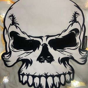 Puede incluir: Un parche de calavera blanco con detalles en negro. El parche presenta un diseño detallado de una calavera con cuencas de ojos, nariz y boca negras. El parche está bordado con las palabras "MEHRAN EMBROIDERY".
