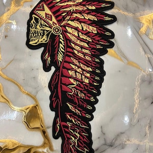 May include: Embroidered patch featuring a skull wearing a Native American headdress. The design incorporates gold and red colors with intricate feather details. The patch has a black border and is likely for jackets or bags.