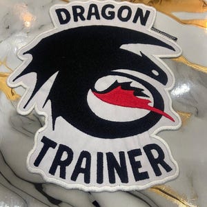 May include: A white embroidered patch with the words "DRAGON" and "TRAINER" in black block letters. The patch features a black dragon silhouette with red accents.