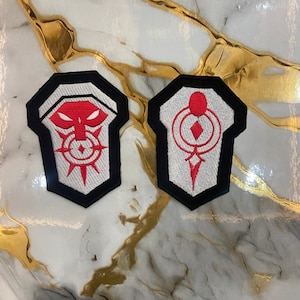 May include: Two embroidered patches with a shield shape. One patch features a red design on a white background with a black border. The other patch has a similar design in red. These patches are likely for clothing or accessories.