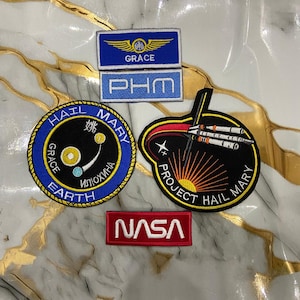 May include: A collection of embroidered patches featuring space-themed designs. Includes patches with the NASA logo, "Project Hail Mary," and "Grace" text. The patches are in various shapes and colors, including blue, black, red, and gold.