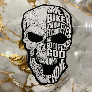 May include: A black and white embroidered patch shaped like a human skull. The left side features a detailed cracked bone texture, while the right side displays the text: SAVE A BIKER OPEN YOUR FUCKING EYES AND GET OFF YOUR GOD DAMN PHONE.