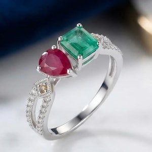 May include: A silver-toned ring featuring a rectangular emerald-cut green gemstone and a pear-shaped red gemstone. The band is accented with small clear stones and a single small brown stone set within a diamond-shaped frame on the side.