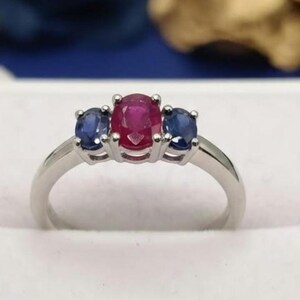 May include: A three-stone ring featuring a central oval-cut red ruby flanked by two smaller oval-cut blue sapphires. The gemstones are set in a polished silver-toned metal band with a prong setting that secures each stone individually.