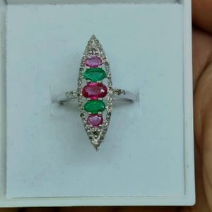 May include: A silver-toned ring featuring a marquise-shaped setting adorned with a vertical row of three oval-cut pink rubies and two oval-cut green emeralds, surrounded by small clear stones along the outer edge of the metal frame.
