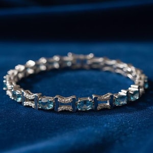 Könnte beinhalten: Ein silberfarbenes Gliederarmband mit abwechselnd oval geschliffenen blauen Edelsteinen und rechteckigen Metallsegmenten, die mit kleinen, klaren weißen Steinen besetzt sind, angeordnet in einer kreisförmigen Form.