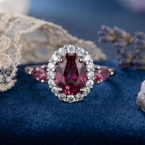 Peut inclure: Une bague ornée d'une pierre centrale ovale rose foncé entourée d'un halo de petites pierres blanches rondes. Deux pierres roses en forme de poire sont serties de chaque côté de la pierre ovale centrale, sur un anneau en métal argenté.
