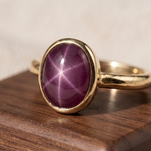 Può includere: Un anello in oro con un cabochon di zaffiro stellato ovale di colore viola intenso come pietra centrale. La gemma mostra un distinto effetto a stella a sei punte sulla sua superficie lucida, incastonato in una semplice fascia d'oro lucida.