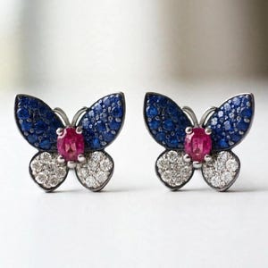 Könnte beinhalten: Ein Paar schmetterlingsförmige Ohrstecker mit dunkelblauem Saphir-Pavé auf den oberen Flügeln, weißem Diamant-Pavé auf den unteren Flügeln und einem zentralen, oval geschliffenen Rubin in einer Fassung aus dunklem Metall.