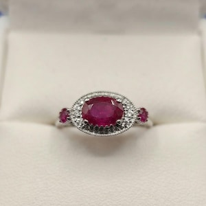 Può includere: Un anello in argento con un rubino centrale rosso intenso dal taglio ovale, circondato da un alone di piccole pietre bianche. La fascia presenta due rubini più piccoli dal taglio rotondo su entrambi i lati dell'incastonatura centrale.