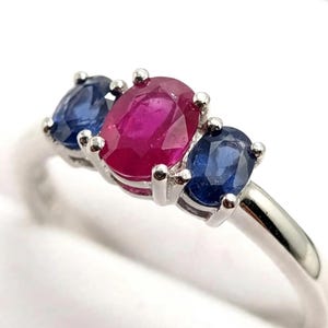 Può includere: Un anello a tre pietre con un rubino rosso centrale dal taglio ovale, affiancato da due zaffiri blu più piccoli dal taglio ovale. Le gemme sono incastonate in una fascia di metallo color argento con montature a quattro griffe.