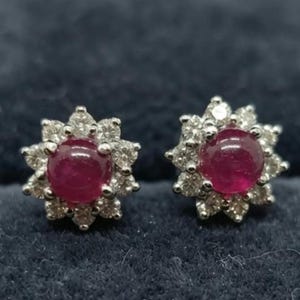 Könnte beinhalten: Ein Paar Ohrstecker mit einem zentralen, rund geschliffenen, tiefroten Rubin-Cabochon, umgeben von einem Kranz aus kleinen, klaren, rund geschliffenen weißen Steinen in einem floralen Muster, gefasst in einen metallischen Rahmen.