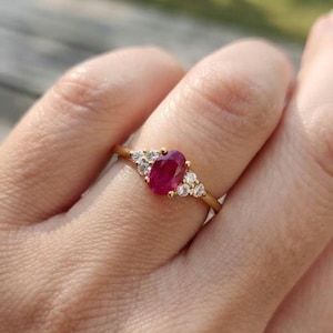 May include: A gold ring featuring a central oval-cut red ruby gemstone flanked by three small round-cut white diamonds on each side. The band is thin and polished, showcasing a classic design with a vibrant red stone and clear sparkling accents.