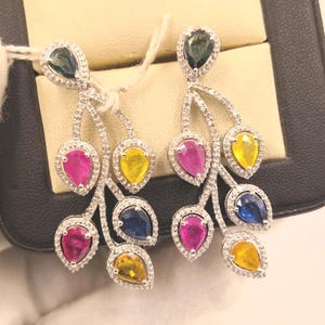 May include: A pair of silver-toned dangle earrings featuring a branch-like design encrusted with small clear stones. Each earring holds multiple pear-shaped gemstones in vibrant shades of pink, yellow, blue, and dark green.