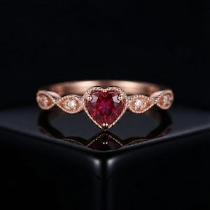 May include: A rose gold ring featuring a central heart-shaped red gemstone. The band is decorated with a series of marquise-shaped settings, each containing a small round white stone, framed by intricate milgrain detailing along the edges.