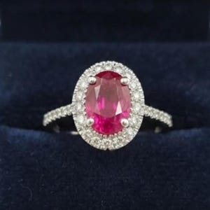 Könnte beinhalten: Ein silberfarbener Ring mit einem großen, oval geschliffenen, leuchtend roten Edelstein in der Mitte. Der zentrale Stein ist von einem Kranz aus kleinen, klaren, rund geschliffenen weißen Steinen umgeben, die auch die Ringschiene zieren.