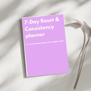Può includere: Un'agenda lilla con il titolo "7-Day Reset & Consistency planner" in testo bianco. Il testo sottostante recita "For overwhelmed people who struggle to start." L'agenda è su una superficie bianca con una pianta secca.