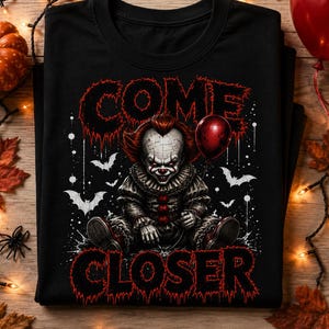 May include: A black t-shirt featuring a graphic of a clown with red hair and makeup, wearing a ruffled collar. The clown holds a red balloon. The text COME CLOSER is printed in a dripping red font above and below the central character illustration.
