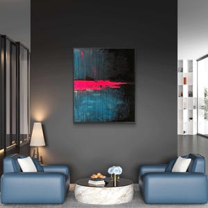 Puede incluir: Pintura abstracta con un fondo oscuro, una vibrante banda horizontal rosa y pinceladas turquesas. La obra de arte enmarcada cuelga de una pared gris oscuro. La habitación cuenta con sillones azules, una mesa de centro redonda y una lámpara de pie.
