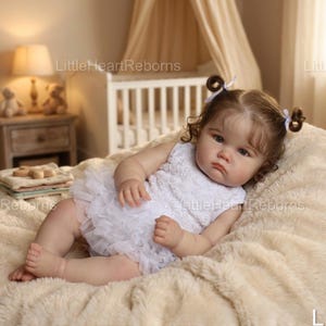 Puede incluir: Una muñeca bebé realista con cabello castaño rizado peinado en coletas, vestida con un vestido blanco sin mangas con una falda con volantes. La muñeca está acostada sobre una manta suave de color crema. La cara de la muñeca tiene una expresión ligeramente triste.