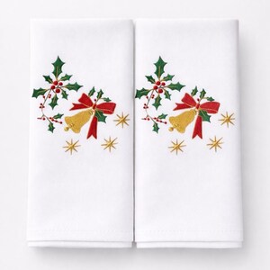 Könnte beinhalten: Zwei weiße Stoffservietten mit gestickten Weihnachtsmotiven. Jede Serviette zeigt eine goldene Glocke mit roter Schleife, grüne Stechpalmenblätter, rote Beeren und goldene Sternakzente. Festliche Weihnachtsdekoration.