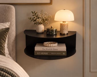 May include: A black semi-circular wall-mounted shelf holding a white vase with flowers, a green lidded jar, a small bowl, and two books titled The Kinfolk Home and AT HOME by Brianna Wiest, illuminated by a white dome-shaped table lamp.