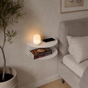 May include: A white, wall-mounted bedside shelf with two curved tiers. A small, glowing lamp, a black smartphone, and a book are on the shelf. A potted plant and a bed with pillows are in the background.