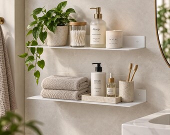 May include: Two white wall-mounted shelves holding bathroom items. The top shelf features a potted plant, a glass jar with cotton swabs, an Aesop hand wash bottle, and a candle. The bottom shelf holds two folded towels, a lotion bottle, a small tray, and a cup with toothbrushes.