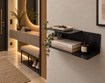 May include: A two-tiered black metal wall shelf holding a beige tray with keys, a small green plant, two books titled The Kinfolk Home and At Home, and a dark glass bottle with black reed diffuser sticks on a light-colored wall.