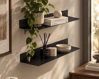 May include: Two matte black metal floating shelves mounted on a wall. The top shelf holds a potted green trailing plant, a round stone sphere, and two stacked books titled The Kinfolk Home and At Home. The bottom shelf holds a reed diffuser.