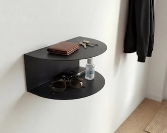 May include: A matte black wall-mounted shelf with a semi-circular design featuring two tiers. The top tier holds a brown leather wallet and a set of metal keys. The bottom tier holds a pair of dark-rimmed sunglasses and a small clear plastic bottle.