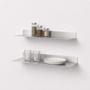 May include: Two brushed metal floating shelves fixed to a white wall. The top shelf holds three glass jars with spices. The bottom shelf displays two textured glasses and two white plates. A minimalist kitchen storage solution.
