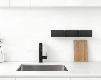 May include: A matte black kitchen faucet with a single handle positioned next to a rectangular stainless steel sink. A long, narrow black wall-mounted shelf is installed above a light brown wooden cutting board.