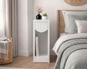 May include: A tall, white, rectangular bedside table featuring a single drawer with a vertical ribbed front panel, an open storage shelf containing books, and a flat top surface holding a black vase with dried flowers and a small potted succulent.