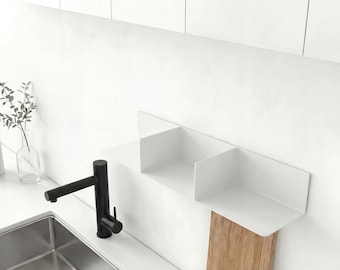 May include: A white metal wall-mounted shelf with two open compartments, positioned above a stainless steel kitchen sink with a black faucet. The shelf features a vertical wooden panel extending downward from the right side section.
