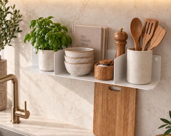 May include: A white metal wall-mounted kitchen shelf holding a potted green plant, three stacked speckled ceramic bowls, a book titled The Kinfolk Table, a wooden pepper mill, a small wooden bowl with salt, and a utensil crock with wooden spoons.