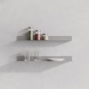 May include: Two horizontal light gray metal wall shelves mounted one above the other. The top shelf holds three glass jars with silver lids containing green herbs, red spices, and brown powder. The bottom shelf holds two ribbed glass tumblers and a white plate.