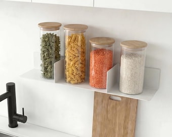 May include: Four clear glass cylindrical storage jars with light wood lids are arranged on a white wall-mounted shelf. The jars contain green pasta, yellow pasta, orange lentils, and white rice. A wooden cutting board hangs below the shelf.